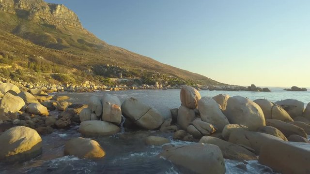 Low altitude drone flight aerial wide shot of massive Eocene primeval coastal sandstone rock formations. Oudekraal Nature Reserve with Victoria Coastal Roadway in golden sunset