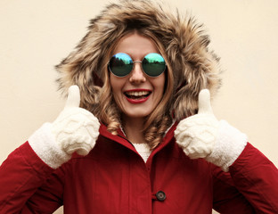Portrait close up happy smiling woman showing thumbs up as like sign wearing red jacket with fur hood and white mittens over background