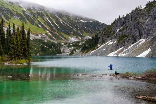 A women hangs out on the side of an alpine lake with her dog and takes in the view in the Coast Mountains around Pemberton, British Columbia, on a rainy summer day.