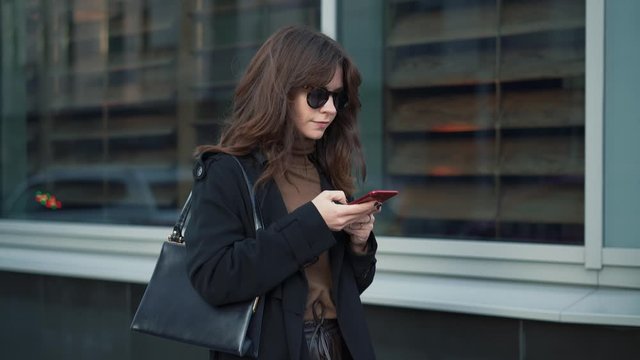 Slow motion of woman in sunglasses with a red phone and a bag on background of shop windows in a big city. Gimbal shot of woman in black coat typing in the phone looking to the road