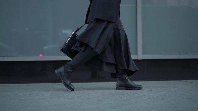 Slow motion gimbal shot of woman dressed in black walking from right to left in black boots on the city pavement on the background of grey shop window glasses