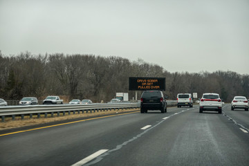Highway view with cars and an SUV passing an electronic traffic warning sign that flashes Drive...