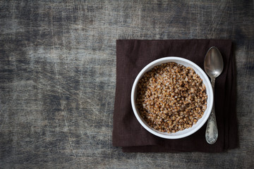 Buckwheat porridge in a white plate and a silver spoon on a dark cloth napkin, placed on a dark wooden background, close up, free space