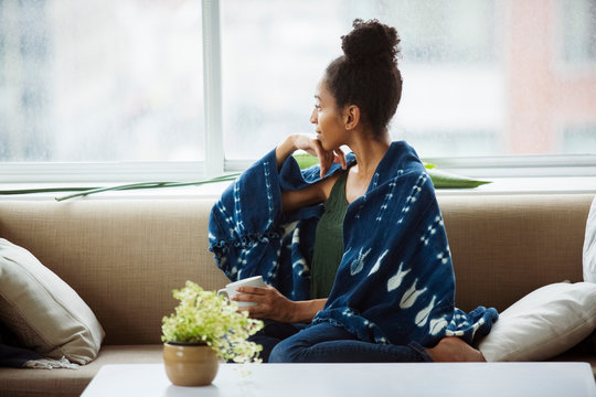 Thoughtful young female wrapped in blanket having coffee on sofa at home