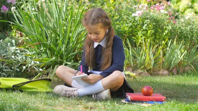 School. Girl schoolgirl sits on the grass and writes in the notebook. 4K UHD