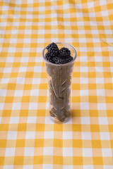 Long glass vase filled with blackberries. Yellow and white checkered textile background.