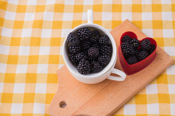 Fototapeta na wymiar Cup and heart-shaped form are filled with blackberries. Wooden kitchen board, textile checkered background.