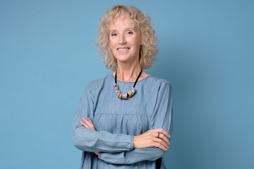 Modern elderly blonde woman standing smiling at the camera with folded arms against blue background
