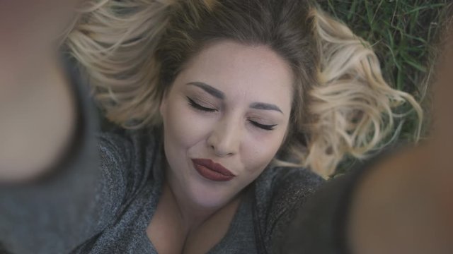 Pretty Woman Smile Close Eyes Lie on Grass Closeup. Attractive Caucasian Girl Photo Pose Face Emotions Expression Top View. Joyful Blonde Lady Laugh Raise Hands Happy Moments Outside Slow Motion 4K
