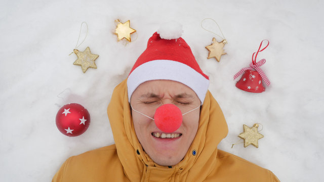 Guy in the image of Santa is lying on the snow around him Christmas toys and shows different emotions. The view from the top, Christmas theme.