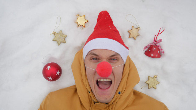 Guy in the image of Santa is lying on the snow around him Christmas toys and shows different emotions. The view from the top, Christmas theme.