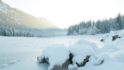 Winterlandschaft: gefrorener See, Wald & Berge, im Vordergrund schneebedeckte Steine