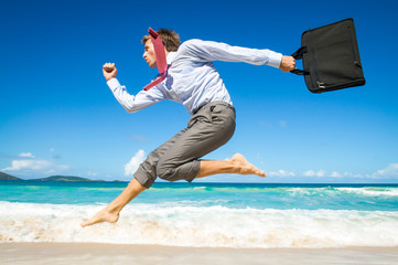 Excited businessman jumping barefoot along a tropical beach swinging his laptop case