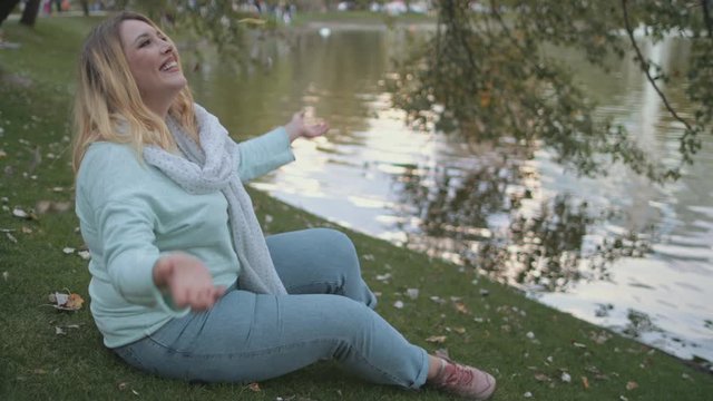 Woman Plus Size Throw Bunch Of Leaves Riverside. Pretty Caucasian Girl Smile Sit by River Bank Outdoors Relaxation Concept. Blonde Hair Lady Relax at City Park Lake Water Surface Slow Motion 4K