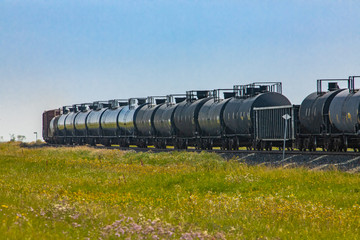 Row of black metallic tank cars of a Canadian freight train. Moving between green fields. Canadian countryside, bright green grass.