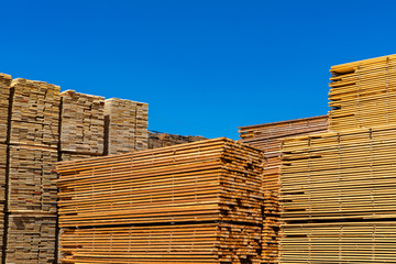 Pallets of treated pine planks are seen stockpiled in a builder merchants yard. Wooden beams and boards beneath a blue sky with copy space.