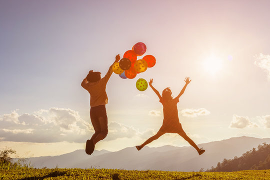 Two children jumping playing with colored balloons at sunset.