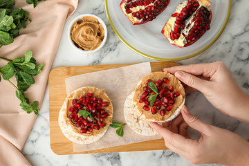 Woman holding puffed rice cake with peanut butter and pomegranate seeds at white marble table, top...