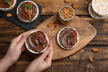 Woman holding puffed rice cake with chocolate spread and grape at wooden table, top view