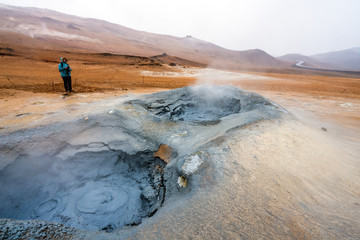 Girl infront of bubbling hot mud pool with mountains and s-shaped road in the background in the...