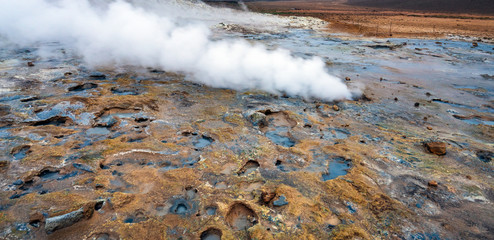Hot sulfuric steam vent spewing sulphur steam in the hot sulfuric and geothermal area of Namaskard in Myvatn/Iceland. Color and mineral rich textured muddy ground infront.