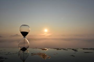 A hourglass (with falling sand) on a wet table with sea of fog and mountain range silhouette in...