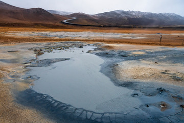 Textured, cracked and patterned ground in the Namaskard/Myvatn area in Icelandic nature. S shaped road and snow covered mountains in the background. Minerals and volcanic energy concept.