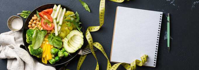 Vegan healthy balanced diet. Vegetarian buddha bowl with blank notebook and measuring tape. Сhickpeas, broccoli, pepper, tomato, spinach, arugula and avocado in plate on dark background. Top view