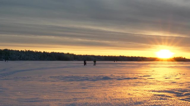 Slider shot of people kick sledding, while others ice fish, on the frozen sea, at the Gulf of Bothnia, at sunset, on a sunny, winter evening, in Ostrobothnia, Finland