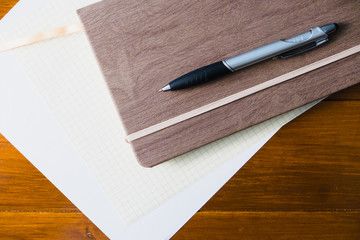 Brown Notebook with white paper and black pen on a rich dark brown wooden tabletop. Journaling and diary writing. Blank page for ideas and taking notes. Writing on wooden grain desktop