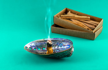 Palo santo, holy wood stick burning / smudging in a natural abalone shell. Bursera graveolens. Palo santo wood sticks in a gift box at background. Isolated on green color background. Copy space.