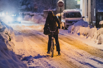 A woman riding a bicycle in the snow on the town street during the early morning hours. Commuting with bike in a cold and snowy weather.