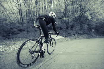 Obraz premium A road cyclist on a black bike is riding in a cold environment. Trees with white patches of snow are seen in the background. Black and white edited image.