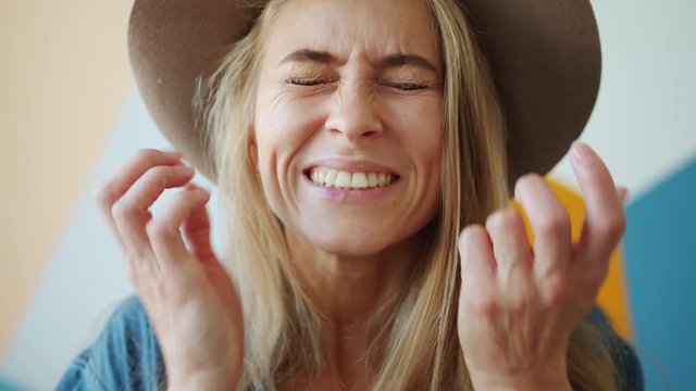 Portrait of cute young blonde making funny faces wearing hat having fun indoors alone on colorful background. Crazy people, grimaces and joy concept.