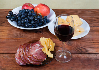 Red wine against cheese, cured pork, fruits on rustic table