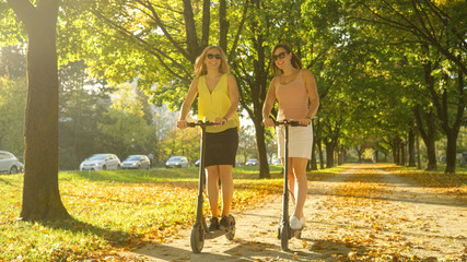 Golden autumn sunbeams shine on cheerful women riding e-scooters around the park