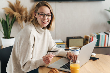 Photo of joyful blonde woman holding credit card and using laptop