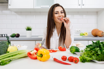 Cheerful young brunette woman tasting bell pepper while cutting it and cooking