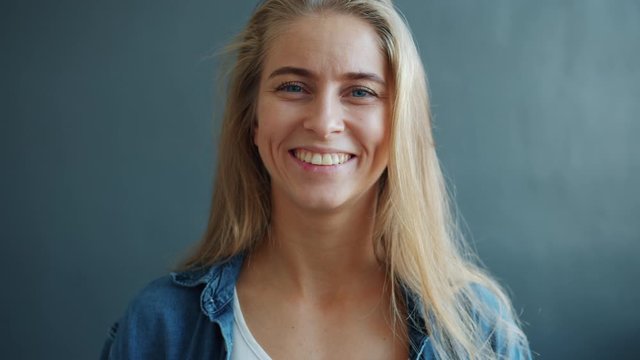 Portrait of cheerful young lady with long blond hair smiling on gray background expressing positive emotions, a cute woman is feeling happy and relaxed.