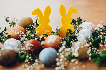Happy Easter. Stylish easter eggs, yellow bunny in nest of spring flowers on rustic wooden table, space for text. Natural dyed easter eggs and rabbit decorations on wooden background
