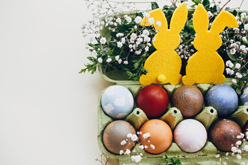 Fototapeta na wymiar Stylish easter eggs in carton tray, yellow bunny in nest of spring flowers on yellow paper, space for text. Natural dyed easter eggs and rabbit decoration. Happy Easter