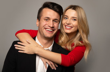 Elegant couple. Close up photo of a charming blonde girl in a red dress, who is hugging her boyfriend in a suit from the back, looking in the camera and smiling.