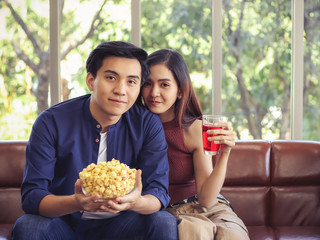 attractive Asian couple in love sitting close together on couch in living room a man holding popcorn bowl  and woman holding red drink.