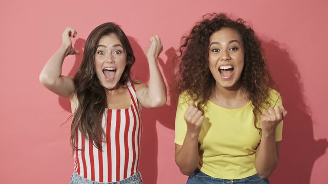 Two cheerful intrigued pretty girls friends experiencing excitement and becoming happy after that while pointing at the camera over pink background
