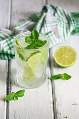 Refreshing cold drink with lime and mint in a glass on a white wooden board table. Traditional summer mojito cocktail. Selective focus.