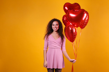 Studio portrait of young woman with dark skin and long curly hair wearing sexy dress over the festive red wall with heart shaped balloon. Close up, isolated background, copy space.