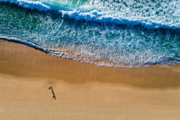 Aerial view of a wave breaking at the shore of the Comporta Beach in Portugal, with a fisherman on the beach.