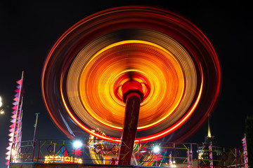 Photo of a carousel at night in a long exposure in an amusement park. The yellow and redder tones of the carousel bulbs merge in motion and give the image a glowing disk effect.
