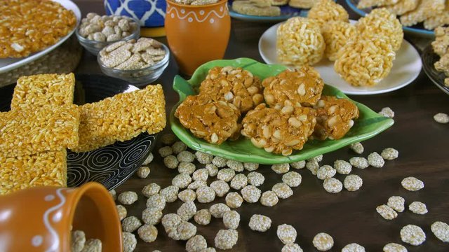 Closeup shot of Lohri items served in ceramic plates and bowls on Lohri festival. A small pot filled with rewri falling on a wooden table surrounded by peanuts chikki  bhujia chikki  murmura ladoo ...