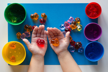 Child hand holding multicolored toys to separating into colorful cups by color on the blue surface